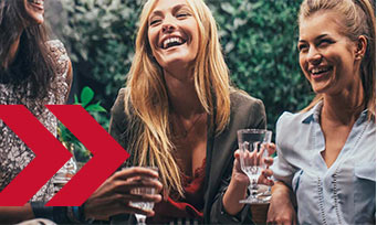 Stock photo of three laughing women holding drinks in garden, red double arrow icon in foreground